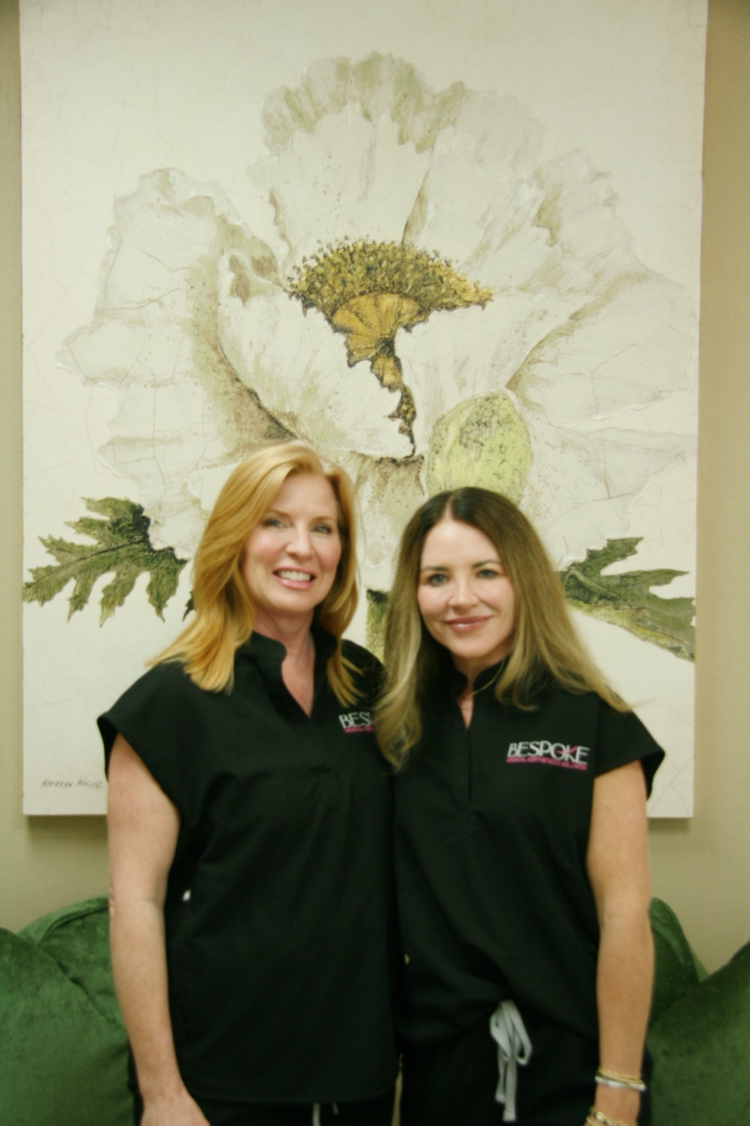 Krista Singleton and Dr. Larissa O&rsquo;Neill, the clinicians leading Bespoke Medical Aesthetics & Wellness, standing together in the sage-walled waiting area beneath a large poppy painting